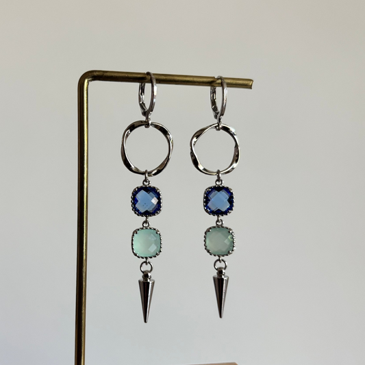 Zilverkleurige oorbellen 'Melly Blauw' op een houten display. De oorbellen bestaan uit een gedraaide cirkel, een donkerblauwe steen, een lichtblauwe steen en een zilveren kegel.