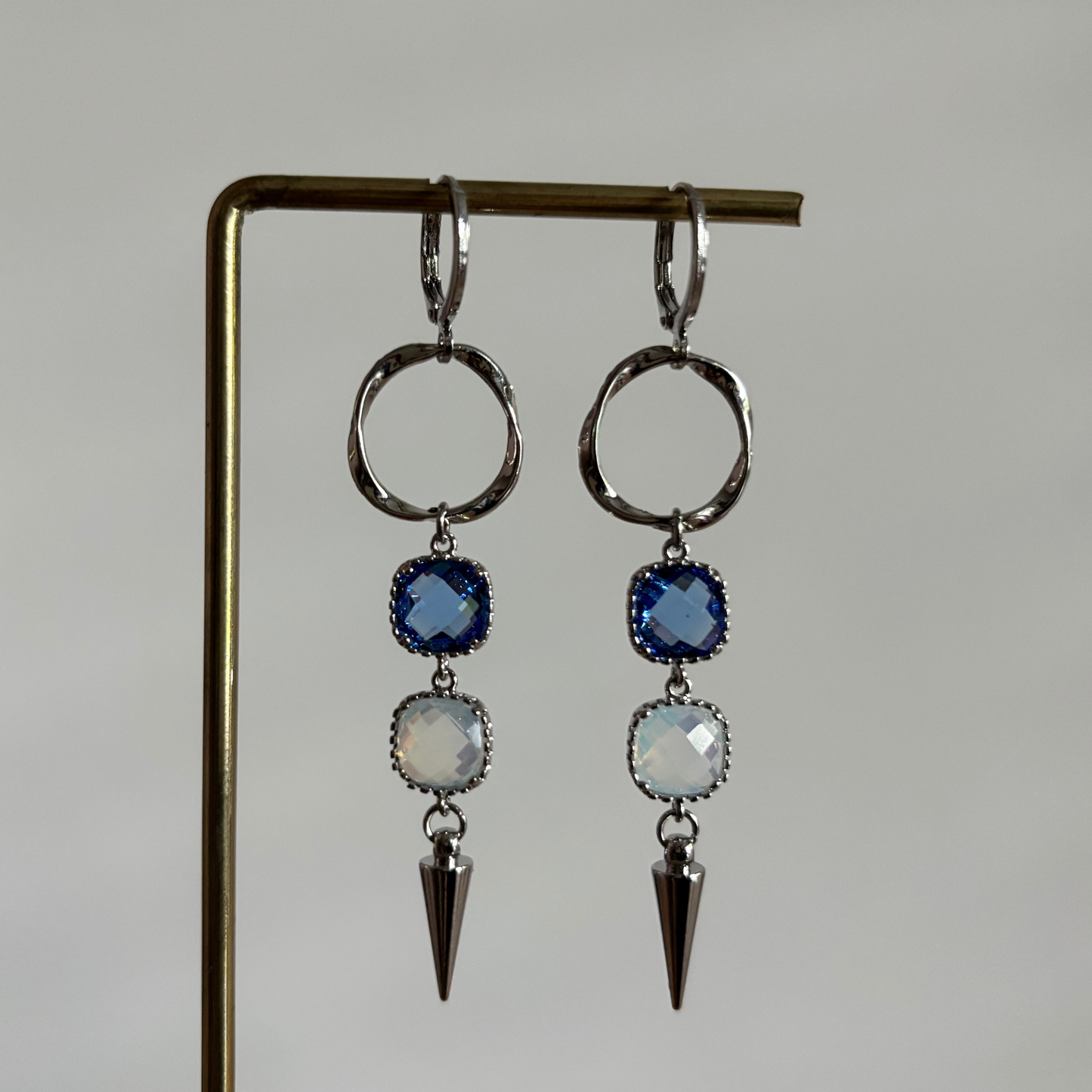 Zilverkleurige oorbellen 'Melly Blauw' op een houten display. De oorbellen bestaan uit een gedraaide cirkel, een donkerblauwe steen, een opal steen en een zilveren kegel.