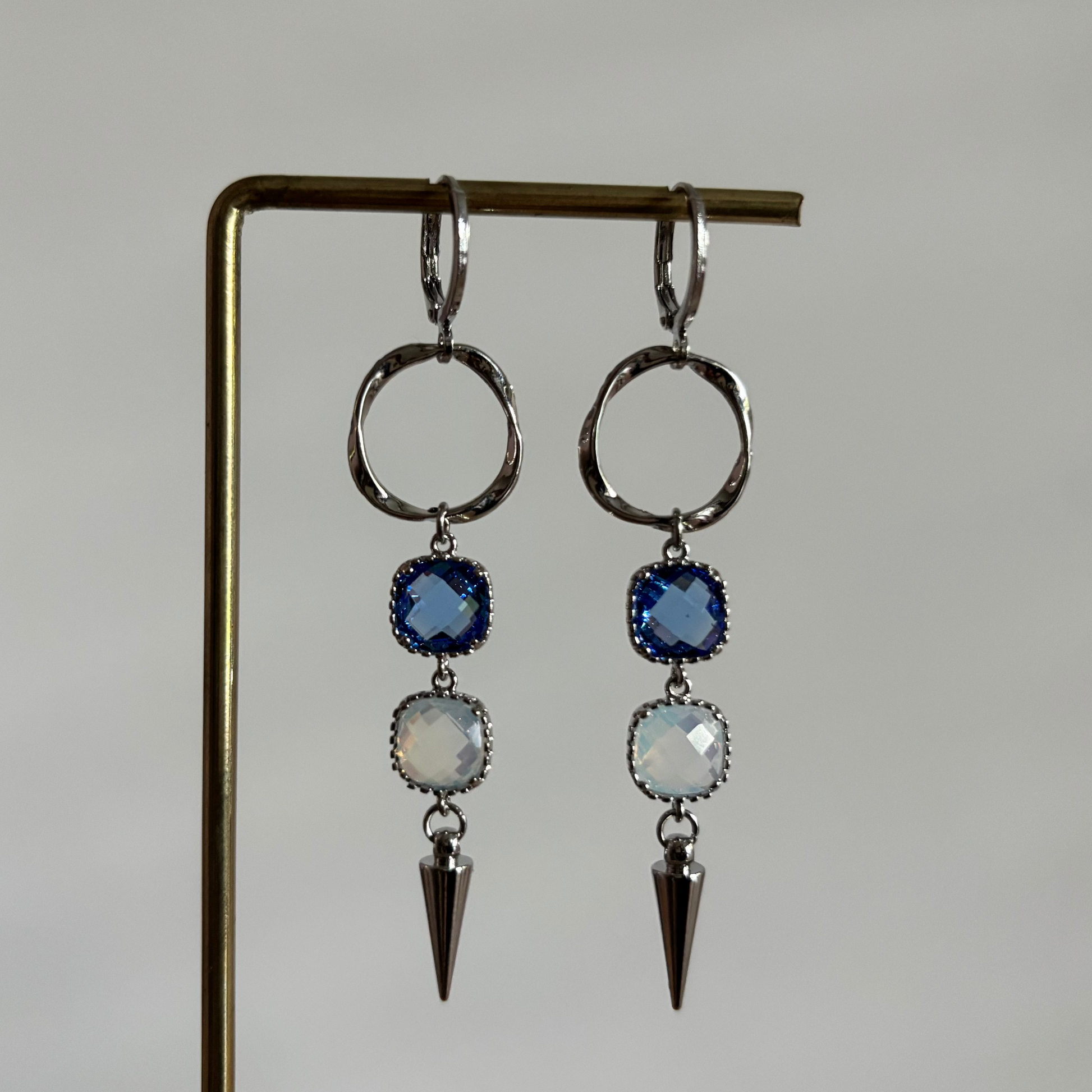 Zilverkleurige oorbellen 'Melly Blauw' op een houten display. De oorbellen bestaan uit een gedraaide cirkel, een donkerblauwe steen, een opal steen en een zilveren kegel.