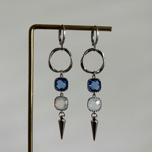 Zilverkleurige oorbellen 'Melly Blauw' op een houten display. De oorbellen bestaan uit een gedraaide cirkel, een donkerblauwe steen, een opal steen en een zilveren kegel.