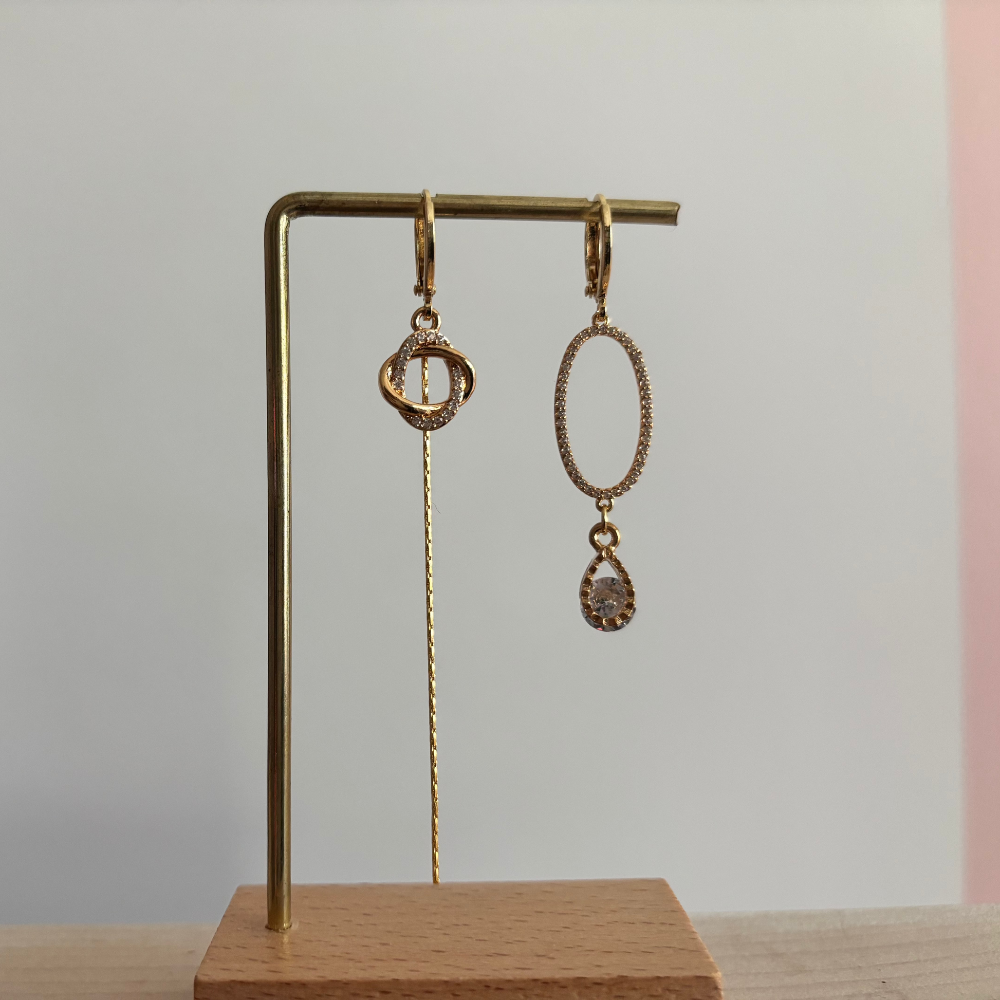 Asymmetrische goudkleurige oorbellen Merle aan een rekje. Links een hanger van twee verstrengelde ringetjes met een lang kettinkje; rechts een grote ovale zirkonia ring met een heldere druppelsteen.