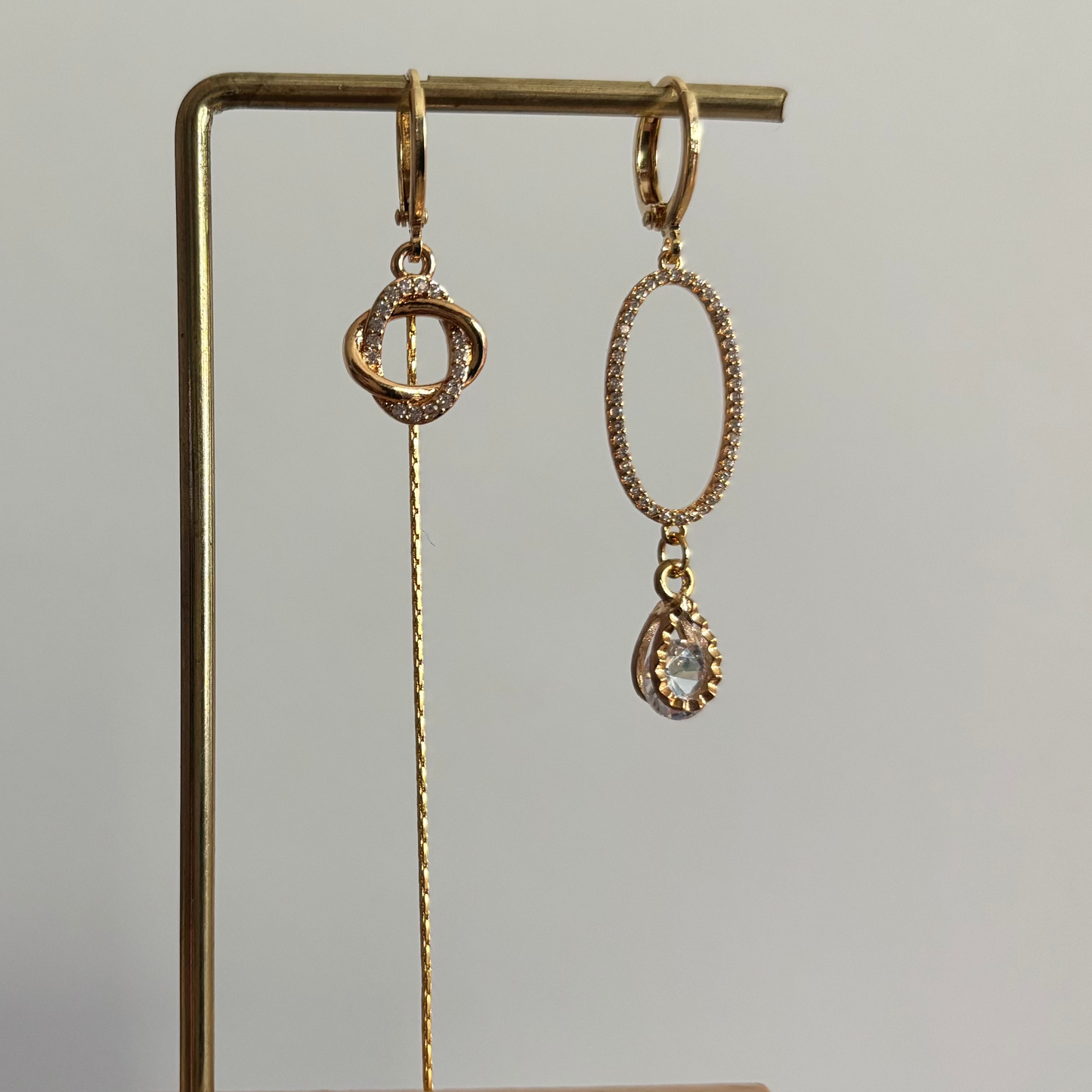 Asymmetrische goudkleurige oorbellen Merle aan een rekje. Links een hanger van twee verstrengelde ringetjes met een lang kettinkje; rechts een grote ovale zirkonia ring met een heldere druppelsteen.