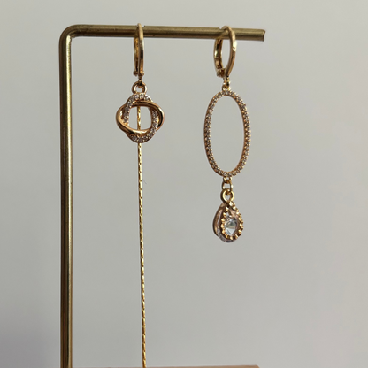 Asymmetrische goudkleurige oorbellen Merle aan een rekje. Links een hanger van twee verstrengelde ringetjes met een lang kettinkje; rechts een grote ovale zirkonia ring met een heldere druppelsteen.