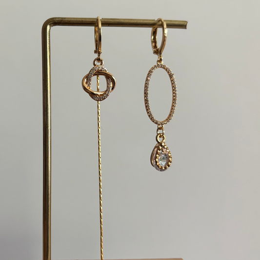 Asymmetrische goudkleurige oorbellen Merle aan een rekje. Links een hanger van twee verstrengelde ringetjes met een lang kettinkje; rechts een grote ovale zirkonia ring met een heldere druppelsteen.