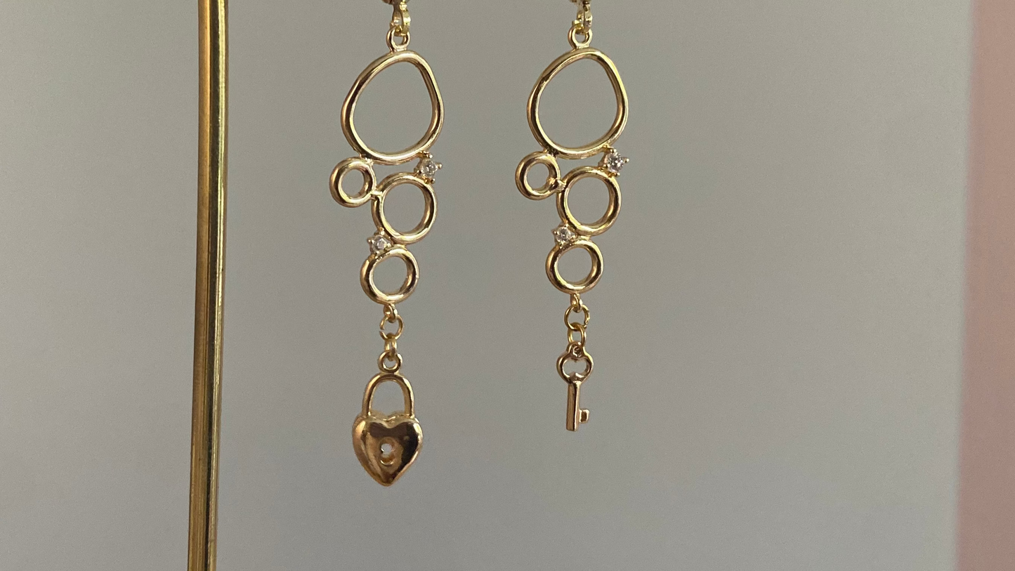 Asymmetrische 14k vergulde oorbellen met cirkeltjeshanger met zirkonia inleg, één met sleuteltje en één met slotje.