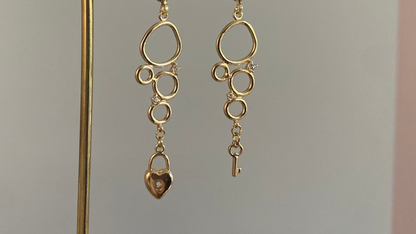 Asymmetrische 14k vergulde oorbellen met cirkeltjeshanger met zirkonia inleg, één met sleuteltje en één met slotje.