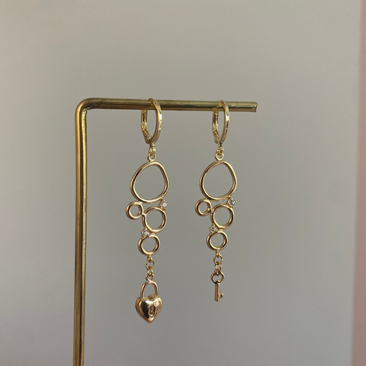 Asymmetrische 14k vergulde oorbellen met cirkeltjeshanger met zirkonia inleg, één met sleuteltje en één met slotje.