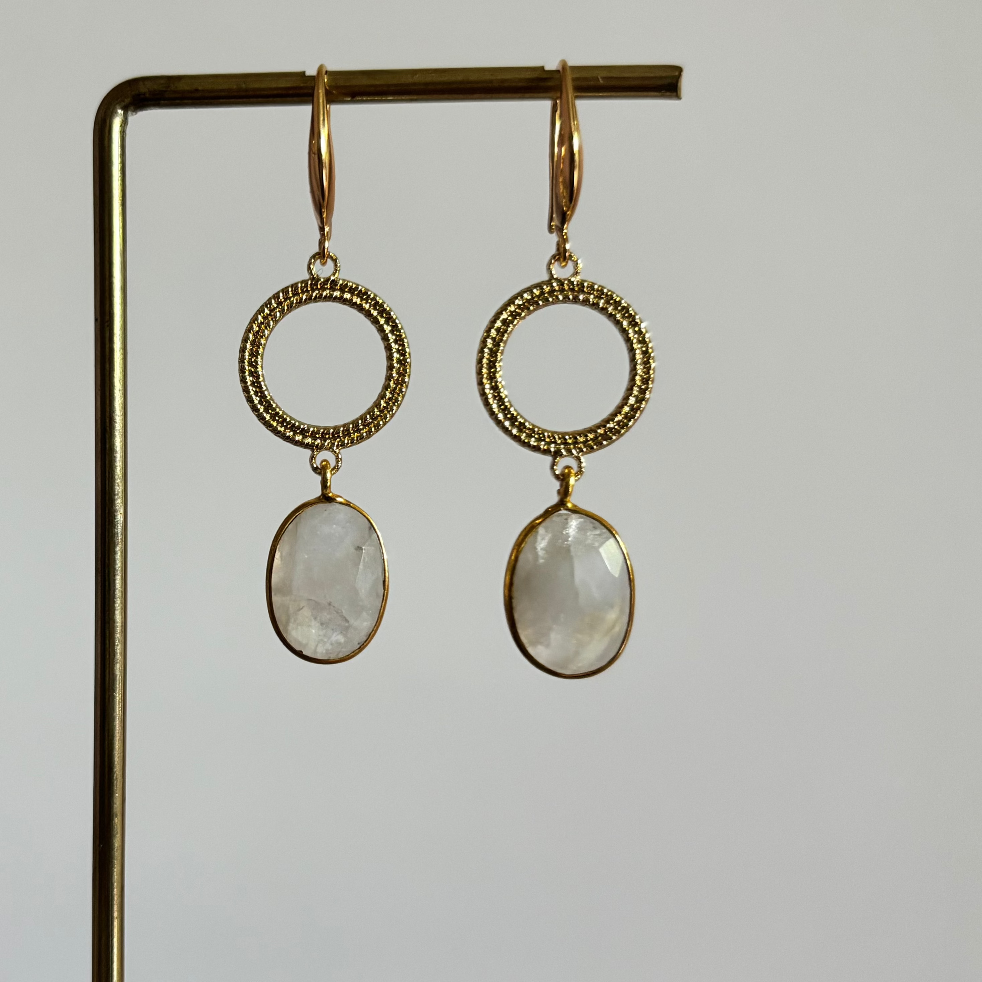 Symmetrische goudkleurige oorbellen Mir Maansteen aan een rekje, voorzien van een ronde open ring met textuur en een hangende, ovaal facetgeslepen witte maansteen.