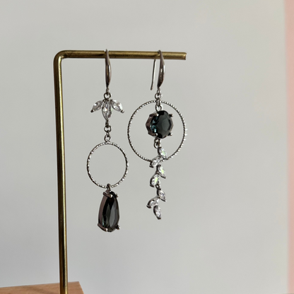 Asymmetrische zilverkleurige oorbellen Mismatch Koosje Grijs aan een rekje. Links een strass-bloemetje met een gedraaide ring en donkergrijze druppel; rechts een grote gedraaide ring met een ronde donkergrijze steen en een hangende strass-rank.