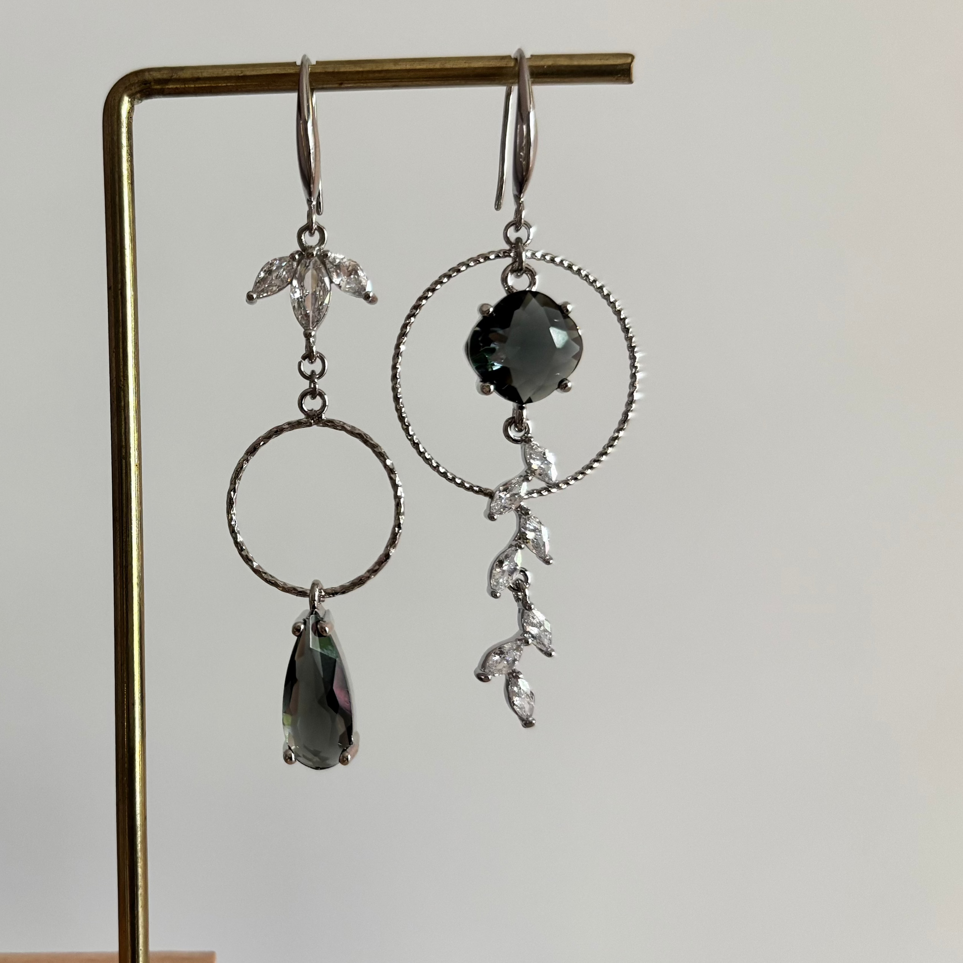 Asymmetrische zilverkleurige oorbellen Mismatch Koosje Grijs aan een rekje. Links een strass-bloemetje met een gedraaide ring en donkergrijze druppel; rechts een grote gedraaide ring met een ronde donkergrijze steen en een hangende strass-rank.