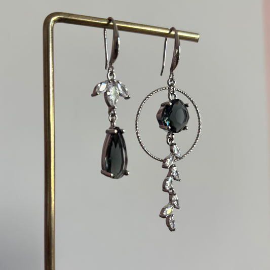 Asymmetrische zilverkleurige oorbellen Mismatch Koosje Grijs aan een rekje. Links een strass-bloemetje met een gedraaide ring en donkergrijze druppel; rechts een grote gedraaide ring met een ronde donkergrijze steen en een hangende strass-rank.