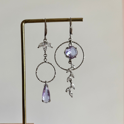 Asymmetrische zilverkleurige oorbellen Mismatch Koosje Lila aan een rekje. Links een strass-bloemetje met een gedraaide ring en lila druppel; rechts een grote gedraaide ring met een ronde lila steen und een hangende strass-rank.