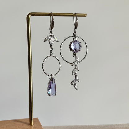 Asymmetrische zilverkleurige oorbellen Mismatch Koosje Lila aan een rekje. Links een strass-bloemetje met een gedraaide ring en lila druppel; rechts een grote gedraaide ring met een ronde lila steen und een hangende strass-rank.