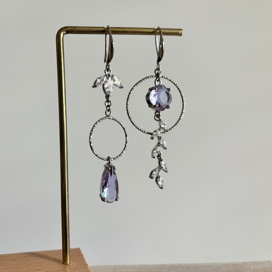 Asymmetrische zilverkleurige oorbellen Mismatch Koosje Lila aan een rekje. Links een strass-bloemetje met een gedraaide ring en lila druppel; rechts een grote gedraaide ring met een ronde lila steen und een hangende strass-rank.