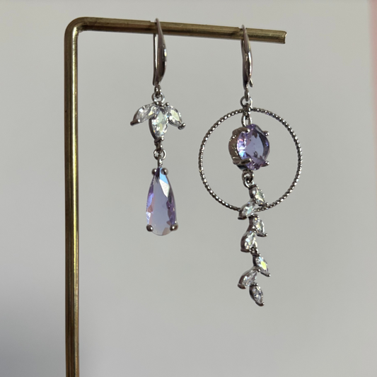 Asymmetrische zilverkleurige oorbellen Mismatch Koosje Lila aan een rekje. Links een strass-bloemetje met een gedraaide ring en lila druppel; rechts een grote gedraaide ring met een ronde lila steen und een hangende strass-rank.