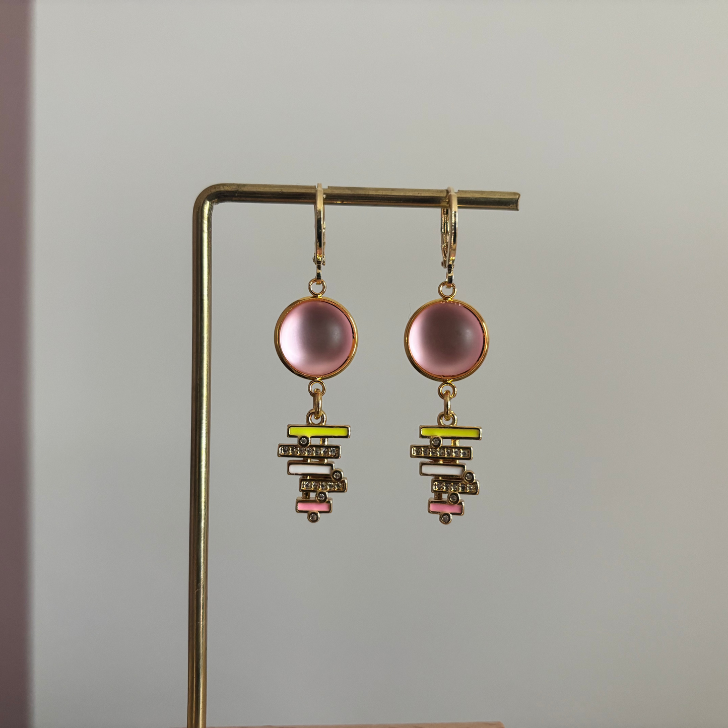 Gouden oorringen met een ronde, lichtroze cabochon steen en een hangende bedel van gestapelde geometrische balkjes in geel, wit en roze emaille, bezet met kleine glimmende steentjes.