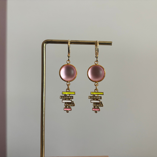 Gouden oorringen met een ronde, lichtroze cabochon steen en een hangende bedel van gestapelde geometrische balkjes in geel, wit en roze emaille, bezet met kleine glimmende steentjes.
