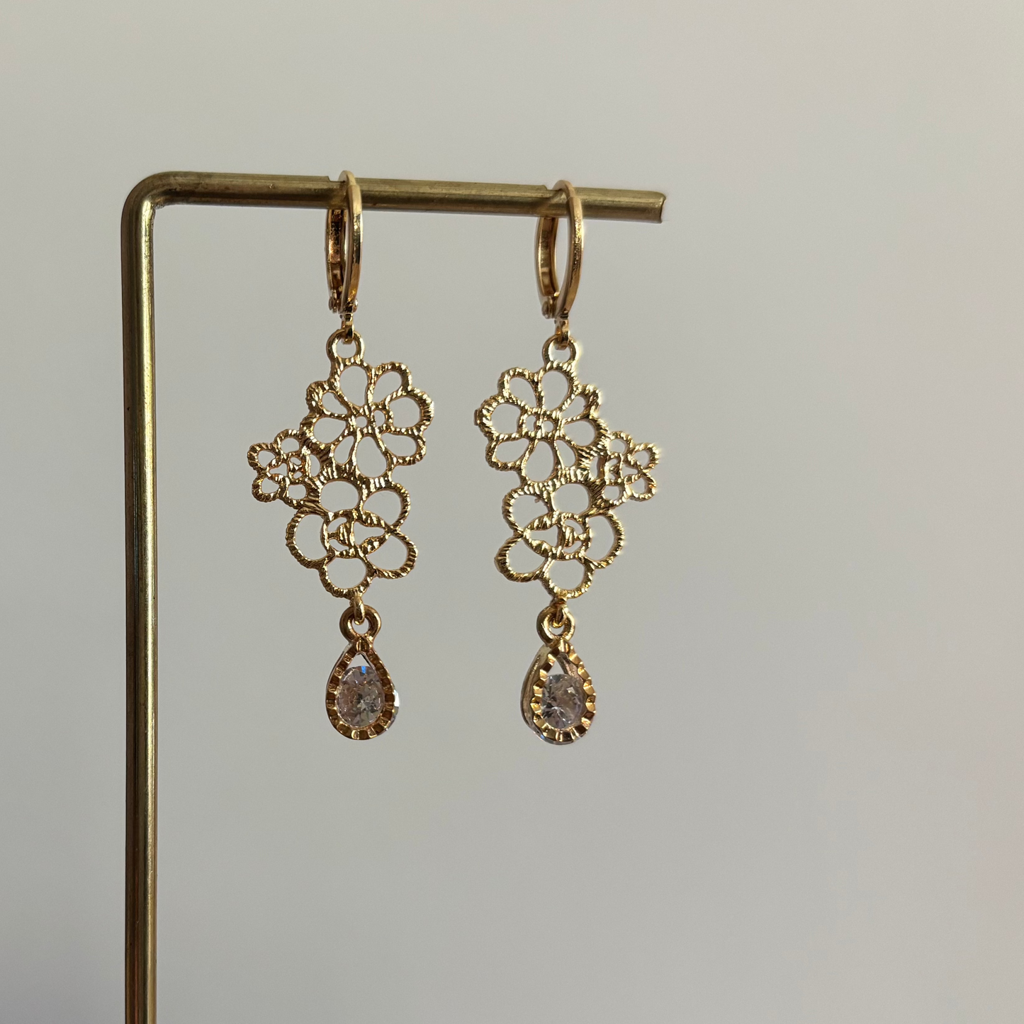 Symmetrische goudkleurige oorbellen Odile aan een rekje, opgebouwd uit een opengewerkte hanger van kleine filigraan bloemetjes en een hangend helder kristal in druppelvorm.