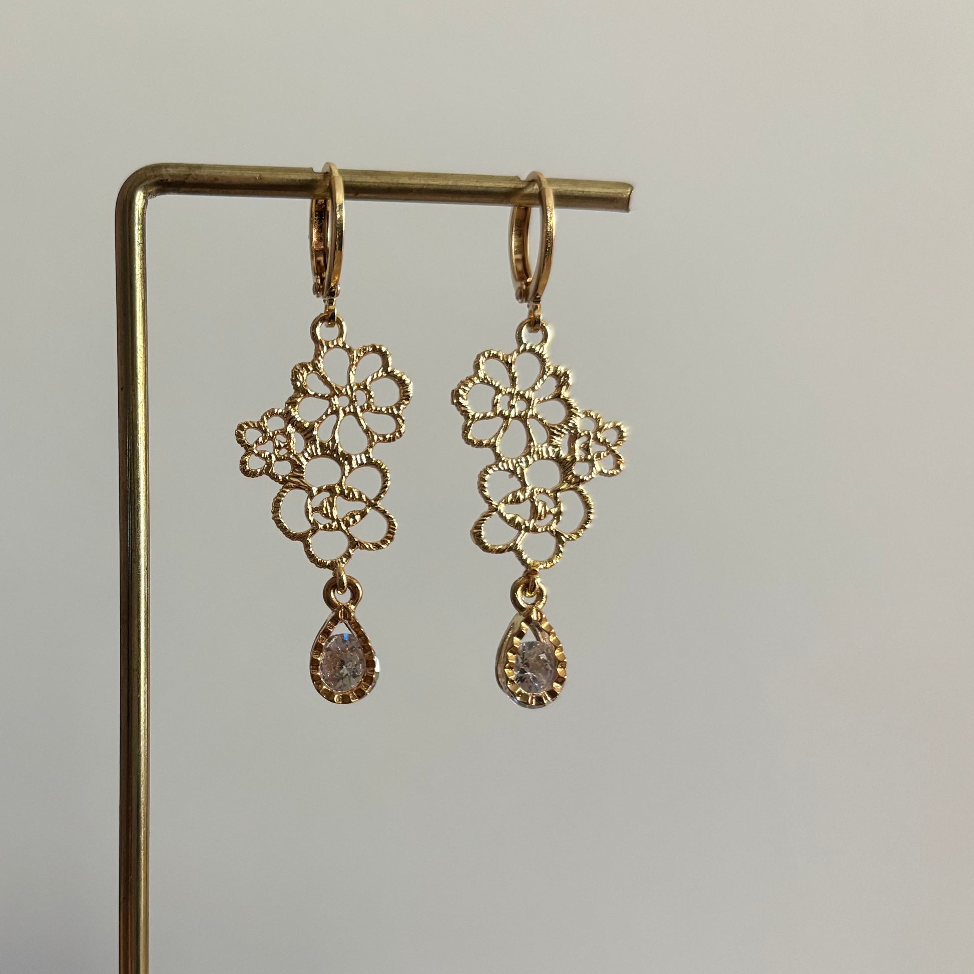 Symmetrische goudkleurige oorbellen Odile aan een rekje, opgebouwd uit een opengewerkte hanger van kleine filigraan bloemetjes en een hangend helder kristal in druppelvorm.