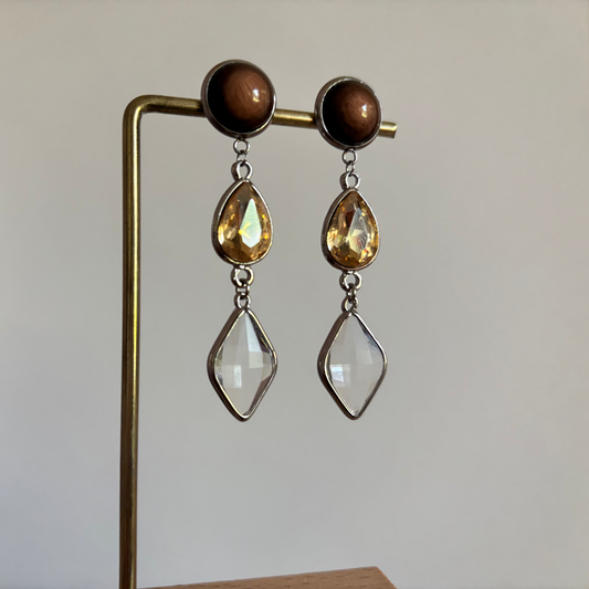 "Zilverkleurige oorhangers 'Oukje Bruin Zilver' met een ronde bruine cabochon knop, een traanvormige amberkleurige glassteen en een heldere ruitvormige hangende steen. Gefotografeerd aan een sieradenstandaard."