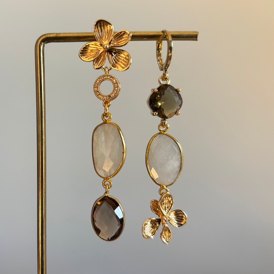 "Lange asymmetrische gouden oorbellen met bloemdetails, melkwitte stenen en rookbruine hangers."