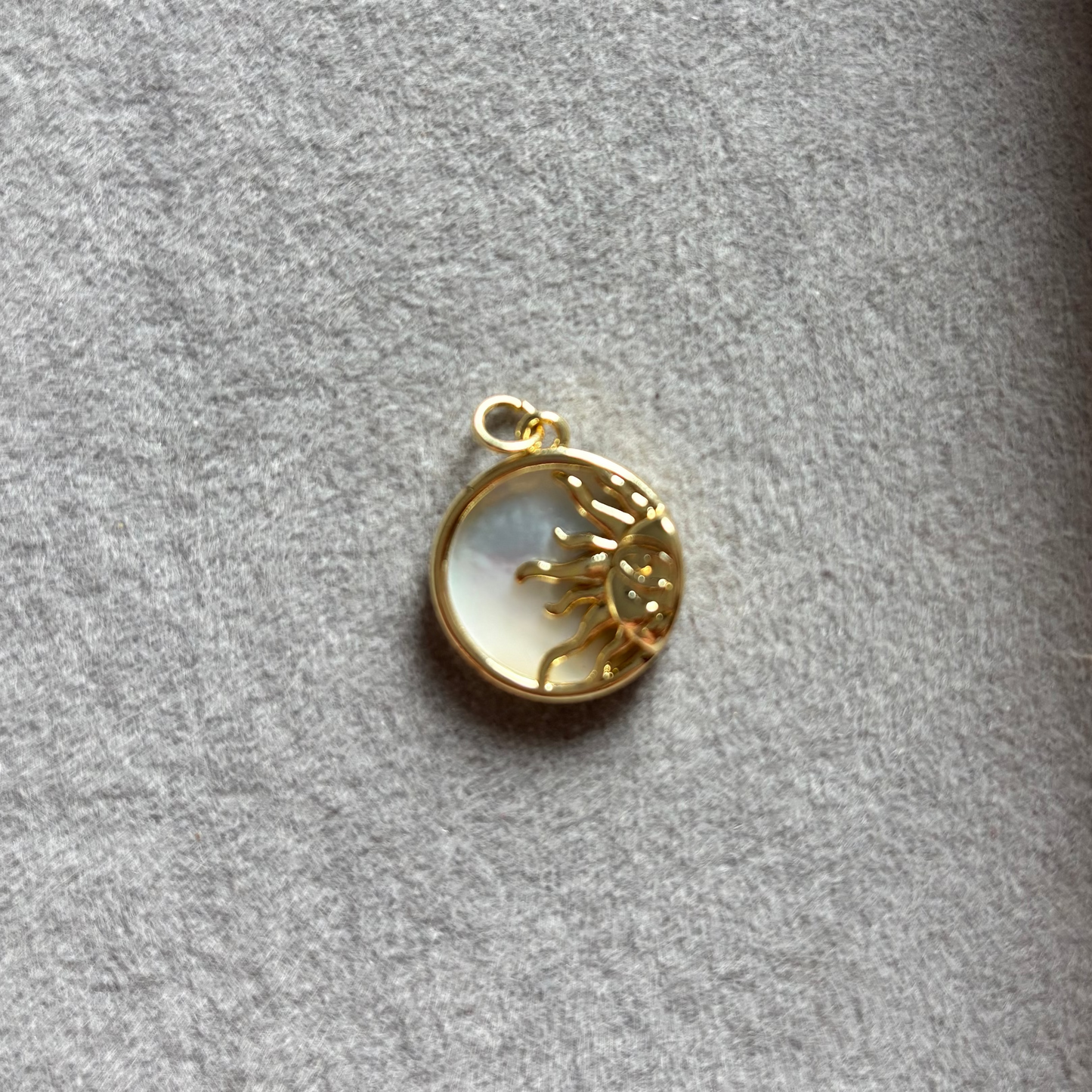 Ronde 14k vergulde bedel met een opengewerkt gouden zonsymbool op een glanzende achtergrond van wit parelmoer, liggend in een grijs sieradendoosje.