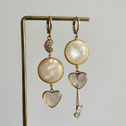 "Gouden mismatched oorbellen set 'Sofie' met ronde parelmoer stenen en hartvormige bedels. De linker oorbel heeft een cabochon met een fijne ketting en strass steentjes; de rechter oorbel heeft een gehamerde gouden schijf en een shell hartje met een gouden rand. Beide oorbellen zijn 14k verguld en hangen aan een gouden display standaard tegen een zachtroze achtergrond."