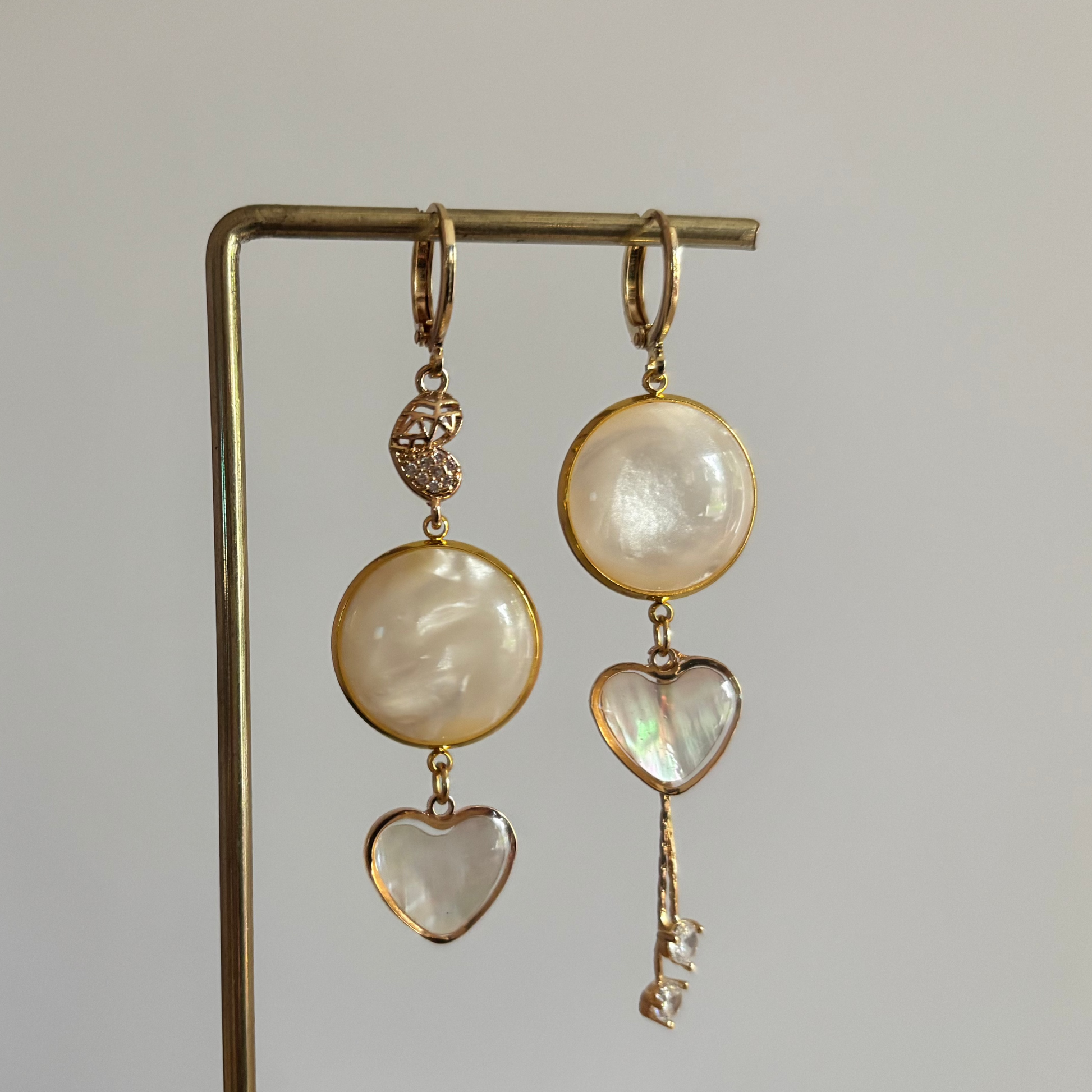 "Gouden mismatched oorbellen set 'Sofie' met ronde parelmoer stenen en hartvormige bedels. De linker oorbel heeft een cabochon met een fijne ketting en strass steentjes; de rechter oorbel heeft een gehamerde gouden schijf en een shell hartje met een gouden rand. Beide oorbellen zijn 14k verguld en hangen aan een gouden display standaard tegen een zachtroze achtergrond."