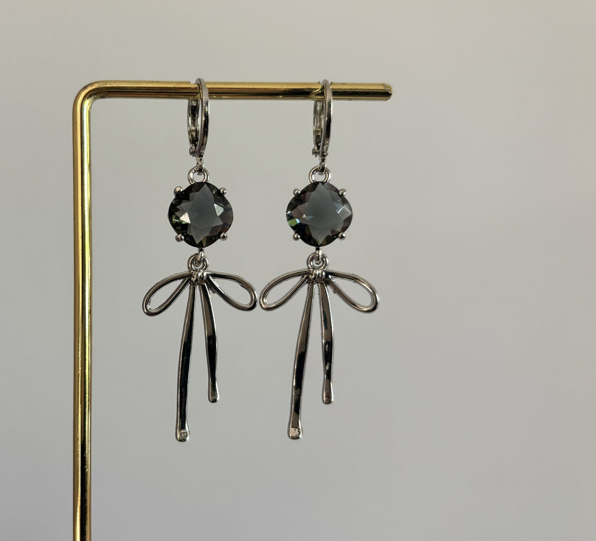Symmetrische zilveren oorbellen van Lellebel hangend aan een gouden standaard. De oorbellen bestaan uit een glanzende zilveren strik met daarboven een grijze glasbedel.