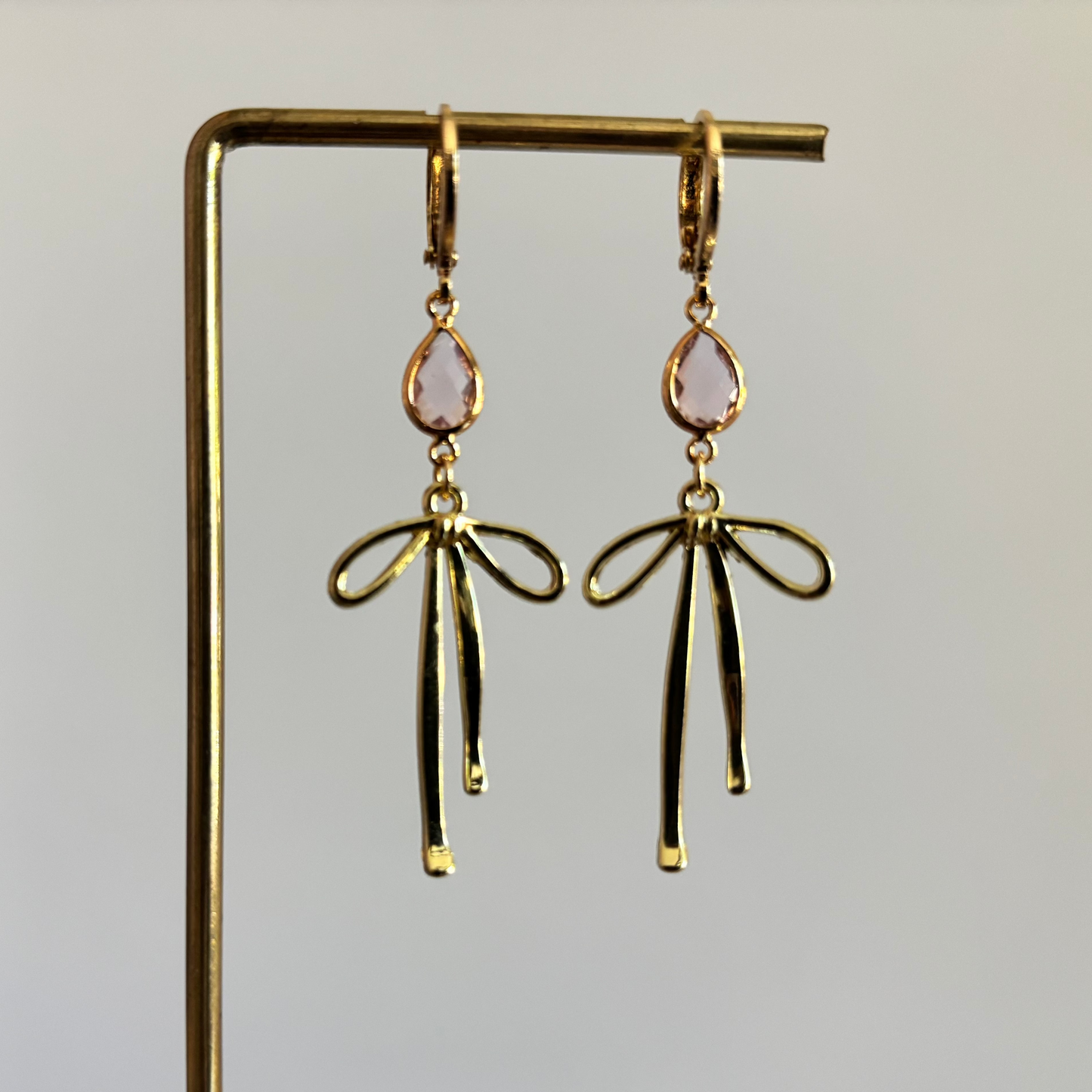 Symmetrische gouden oorbellen van Lellebel op een gouden display. De oorbellen bestaan uit een 14k verguld huggie-ringetje met daaraan een lichtroze, facetgeslepen druppelsteen in een gouden zetting en een elegant gouden strikje daaronder.