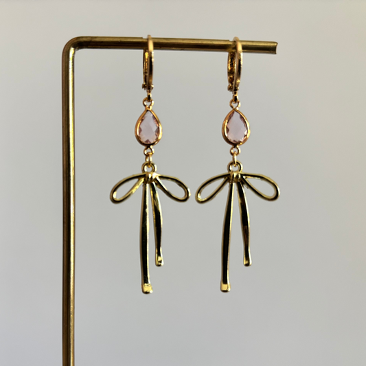 Symmetrische gouden oorbellen van Lellebel op een gouden display. De oorbellen bestaan uit een 14k verguld huggie-ringetje met daaraan een lichtroze, facetgeslepen druppelsteen in een gouden zetting en een elegant gouden strikje daaronder.