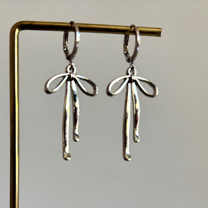 Symmetrische zilveren oorbellen van Lellebel hangend aan een gouden standaard. De oorbellen bestaan uit een verzilverd huggie-ringetje met daaraan een verfijnde, glanzende zilveren strik.