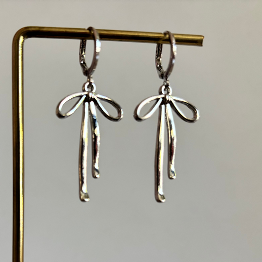 Symmetrische zilveren oorbellen van Lellebel hangend aan een gouden standaard. De oorbellen bestaan uit een verzilverd huggie-ringetje met daaraan een verfijnde, glanzende zilveren strik.