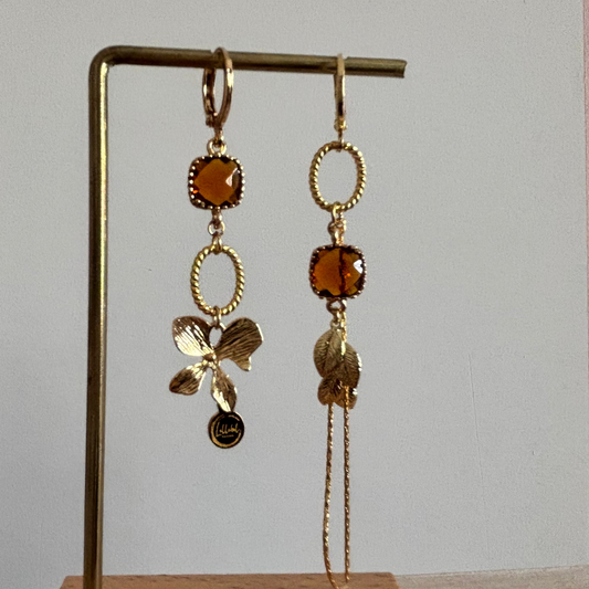 Goudkleurige asymmetrische oorbellen Terri met vierkante amberkleurige kristalstenen, een gouden strikbedel, blaadjes en een fijn kettinkje.