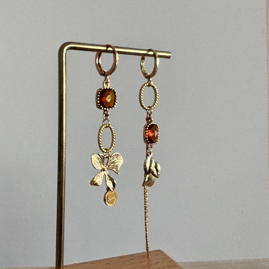Goudkleurige asymmetrische oorbellen Terri met vierkante amberkleurige kristalstenen, een gouden strikbedel, blaadjes en een fijn kettinkje.