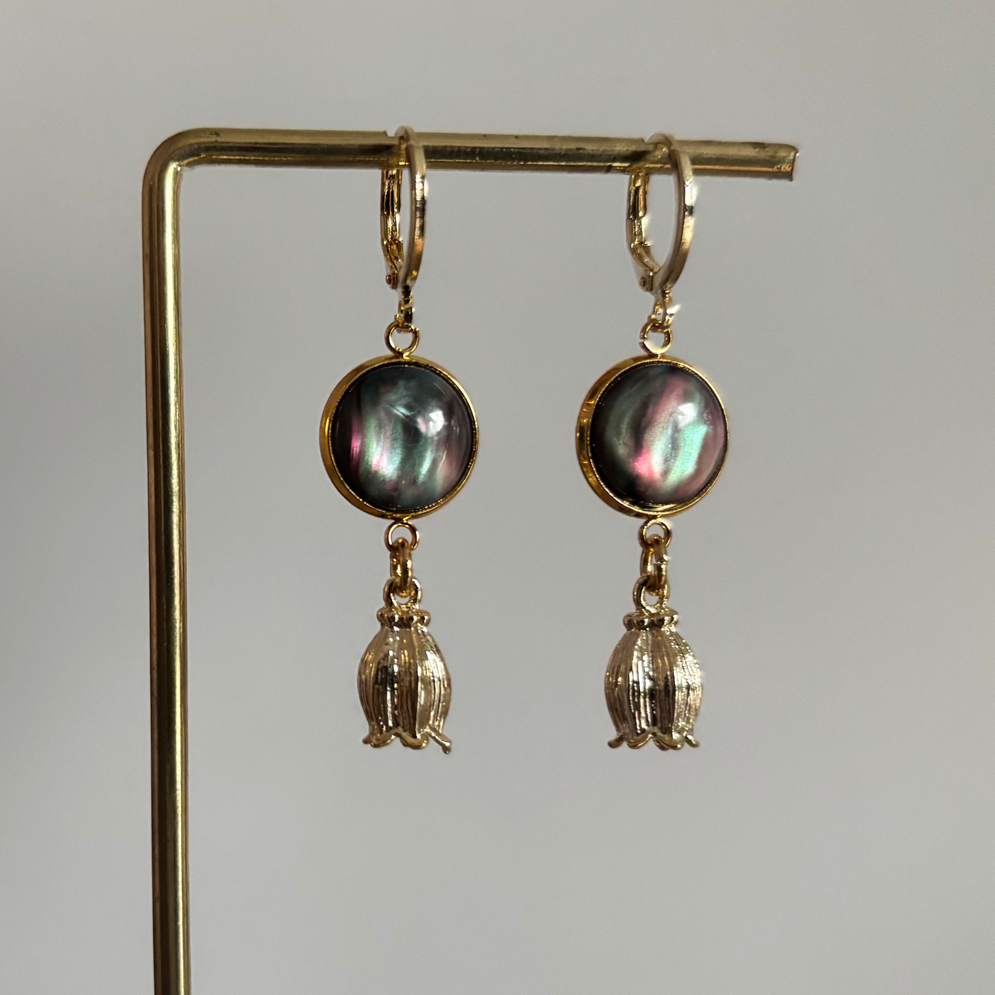 Korte 14k vergulde oorbellen 'Tulp' van Lellebel op een gouden standaard. De oorbellen hebben een ronde, grijze cabochon met een parelmoerglans en een roze gloed, met daaronder een goudkleurige bedel in de vorm van een tulpje.
