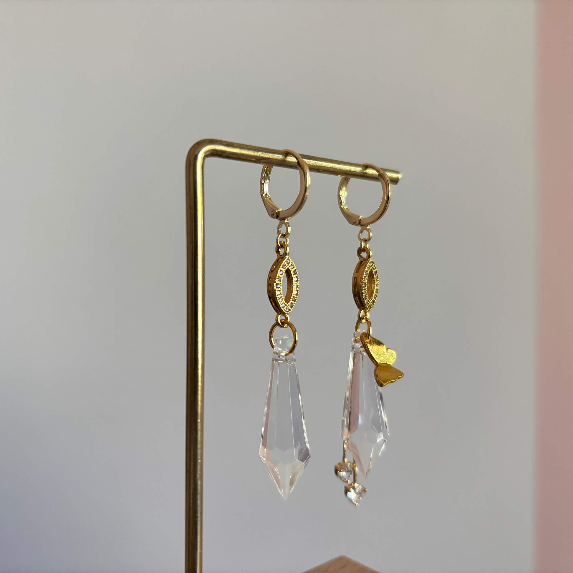 Asymmetrische 14k gouden oorbellen genaamd 'Vlinder' op een display. Links een grote heldere acrylkristal. Rechts dezelfde kristal met een gouden vlinderbedel en twee kleine kristallen. Ringetjes met zirkonia