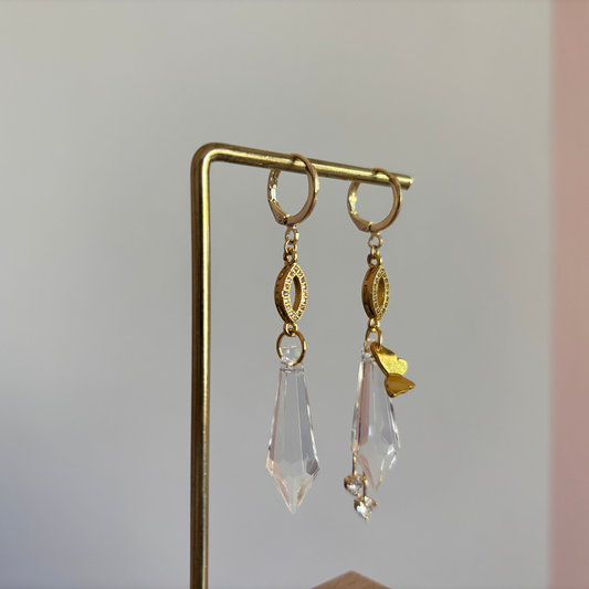 Asymmetrische 14k gouden oorbellen genaamd 'Vlinder' op een display. Links een grote heldere acrylkristal. Rechts dezelfde kristal met een gouden vlinderbedel en twee kleine kristallen. Ringetjes met zirkonia