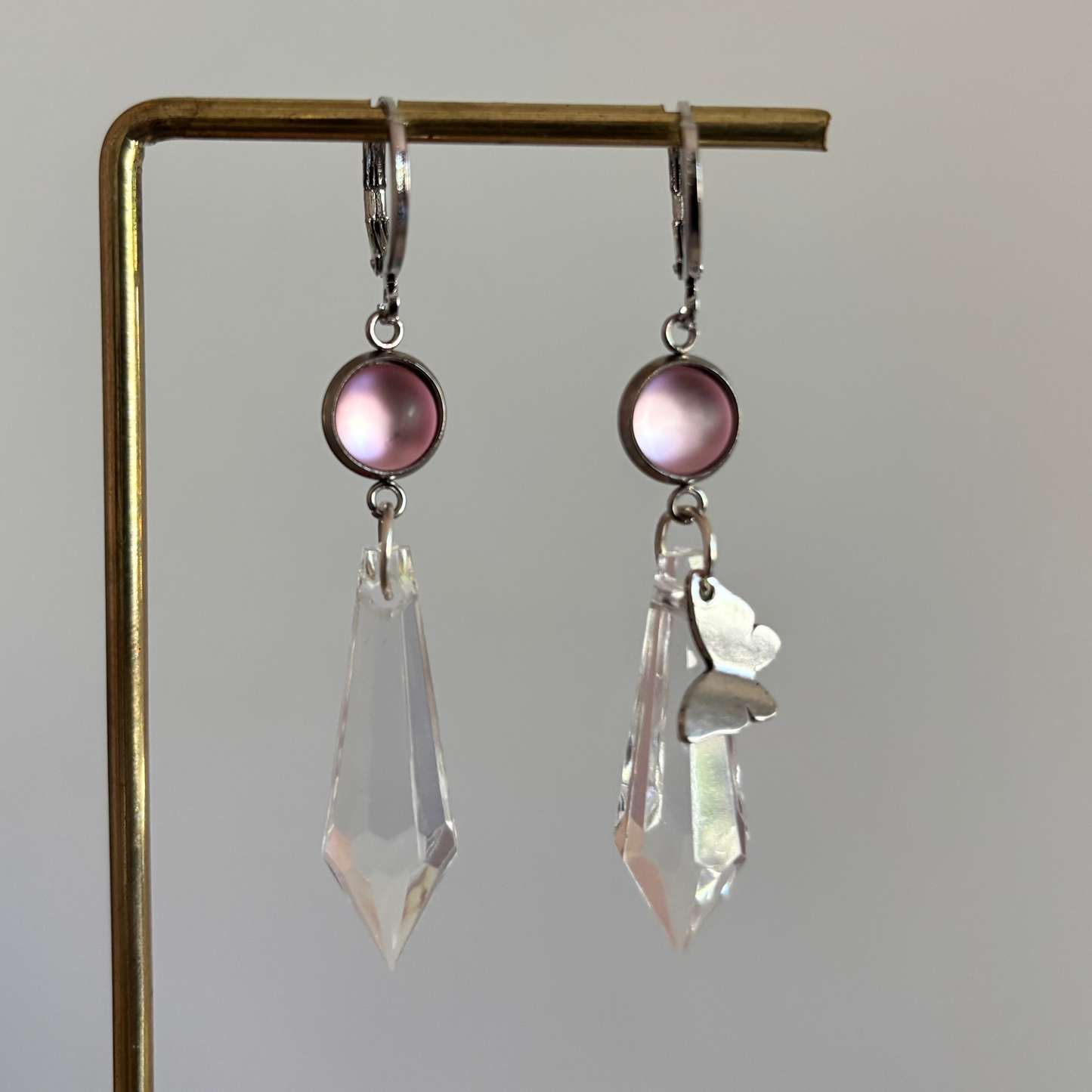 Asymmetrische zilverkleurige oorbellen genaamd 'Vlinder Zilver' op een display. Beiden hebben een ronde roze steen en een grote heldere acrylkristal. Rechts met een zilveren vlinderbedel
