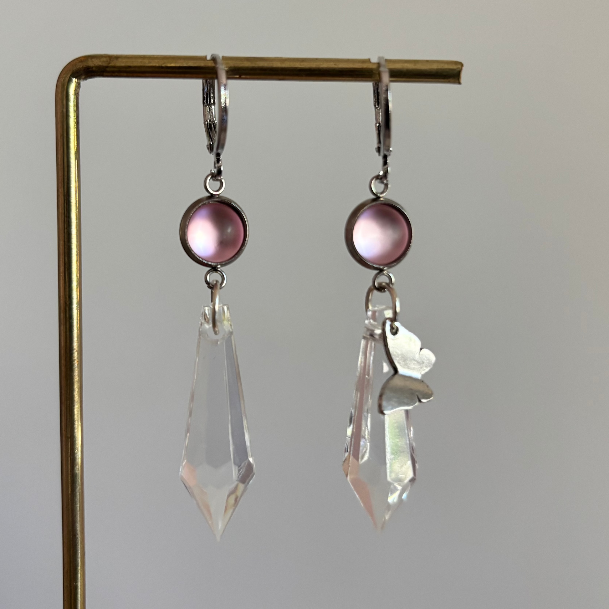 Asymmetrische zilverkleurige oorbellen genaamd 'Vlinder Zilver' op een display. Beiden hebben een ronde roze steen en een grote heldere acrylkristal. Rechts met een zilveren vlinderbedel