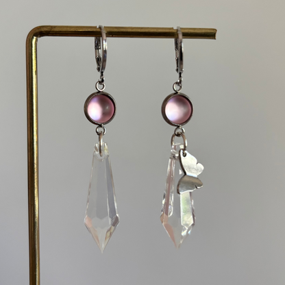 Asymmetrische zilverkleurige oorbellen genaamd 'Vlinder Zilver' op een display. Beiden hebben een ronde roze steen en een grote heldere acrylkristal. Rechts met een zilveren vlinderbedel