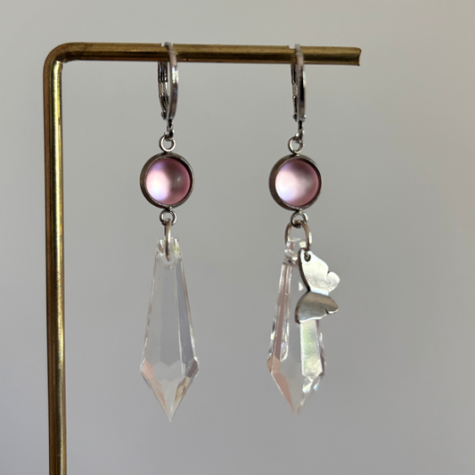 Asymmetrische zilverkleurige oorbellen genaamd 'Vlinder Zilver' op een display. Beiden hebben een ronde roze steen en een grote heldere acrylkristal. Rechts met een zilveren vlinderbedel
