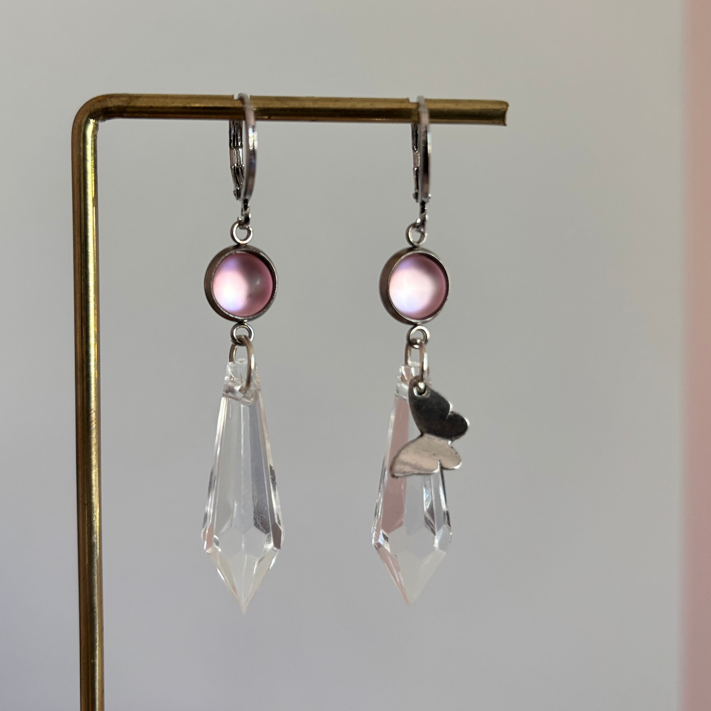 Asymmetrische zilverkleurige oorbellen genaamd 'Vlinder Zilver' op een display. Beiden hebben een ronde roze steen en een grote heldere acrylkristal. Rechts met een zilveren vlinderbedel