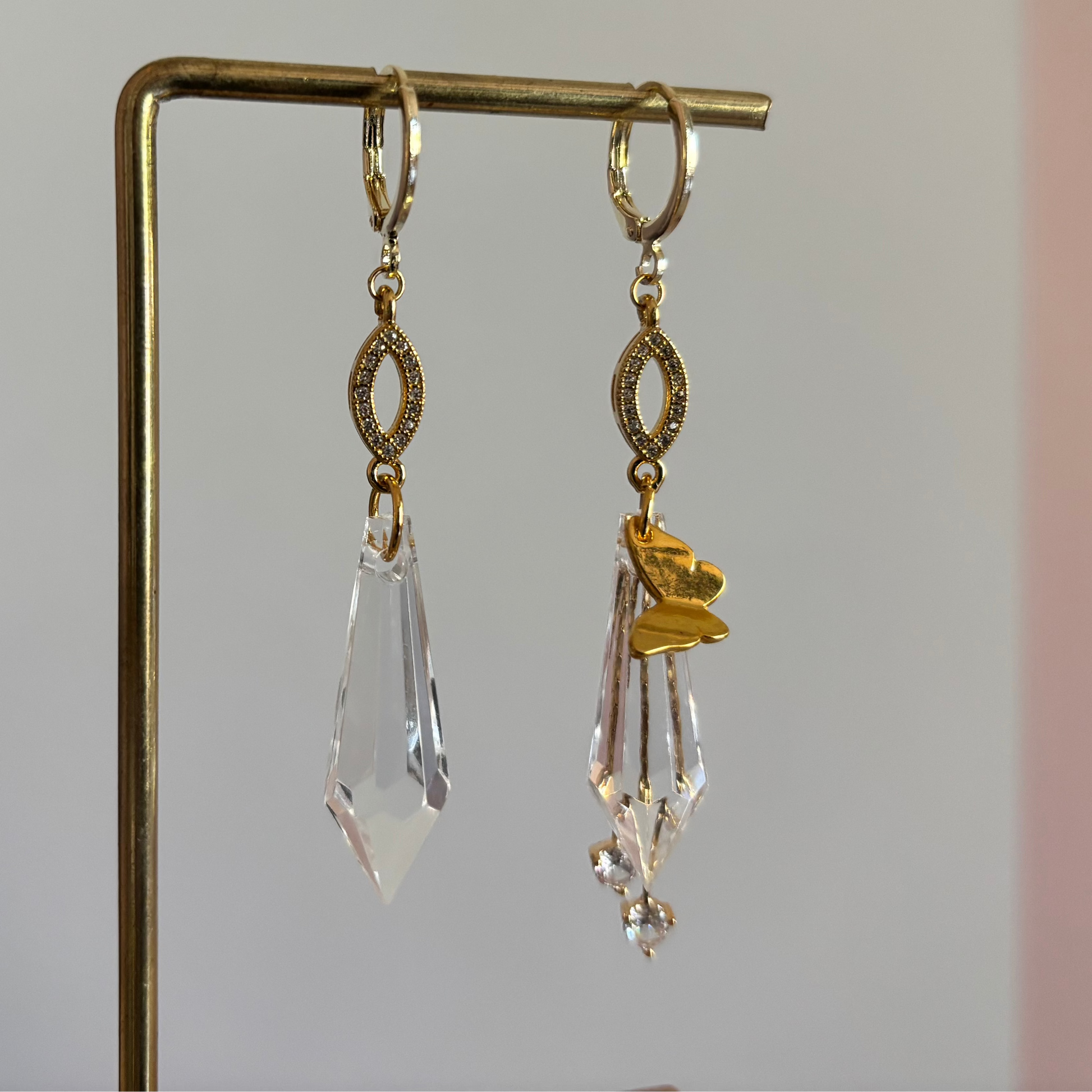 Asymmetrische 14k gouden oorbellen genaamd 'Vlinder' op een display. Links een grote heldere acrylkristal. Rechts dezelfde kristal met een gouden vlinderbedel en twee kleine kristallen. Ringetjes met zirkonia