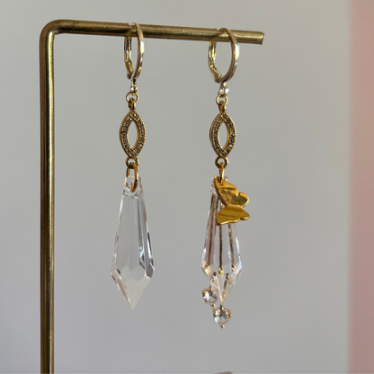 Asymmetrische 14k gouden oorbellen genaamd 'Vlinder' op een display. Links een grote heldere acrylkristal. Rechts dezelfde kristal met een gouden vlinderbedel en twee kleine kristallen. Ringetjes met zirkonia