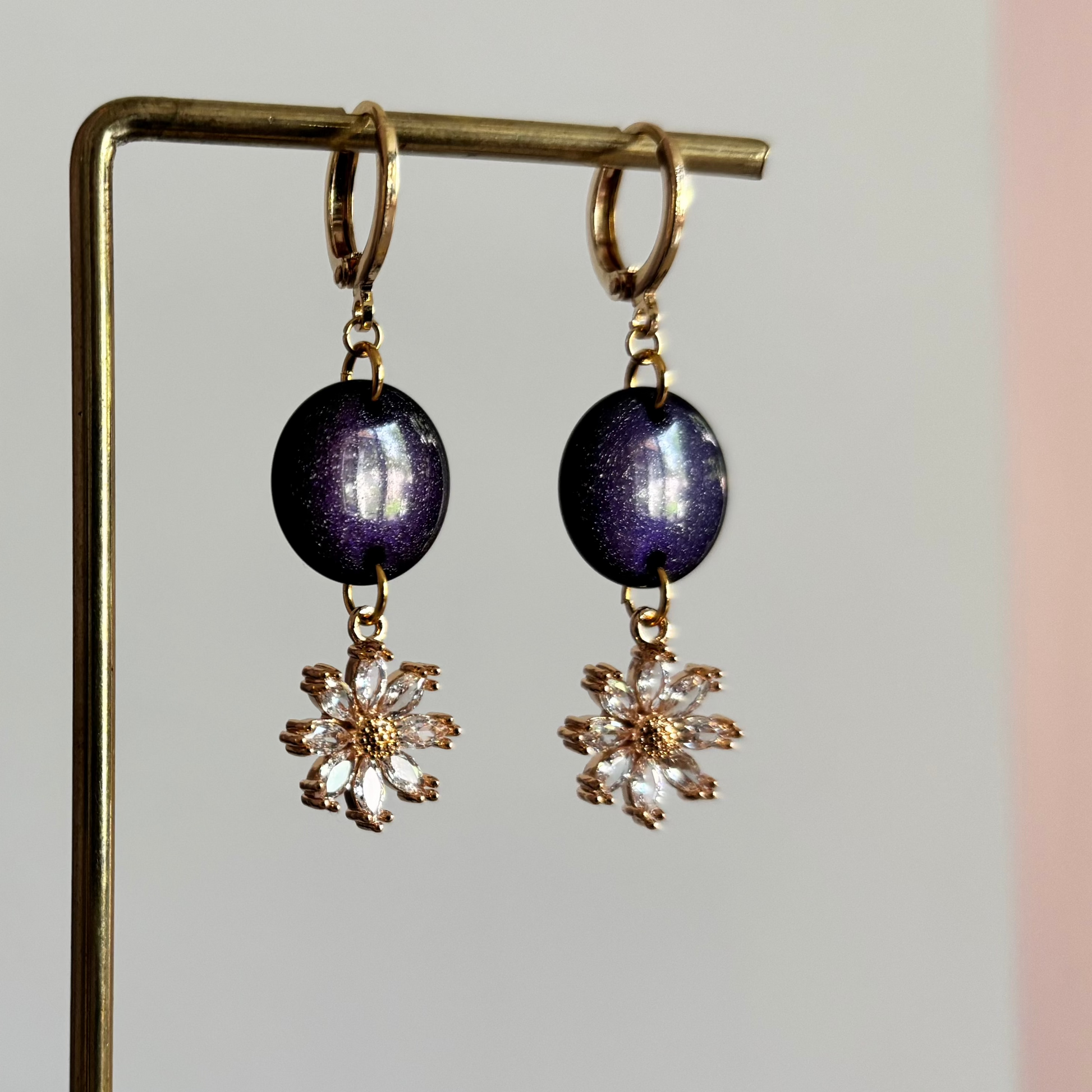 Goudkleurige oorbellen Wendy aan een rekje, voorzien van een ronde, donkerblauwe cabochon met een lichte glitter en daaronder een zirkonia bloem.
