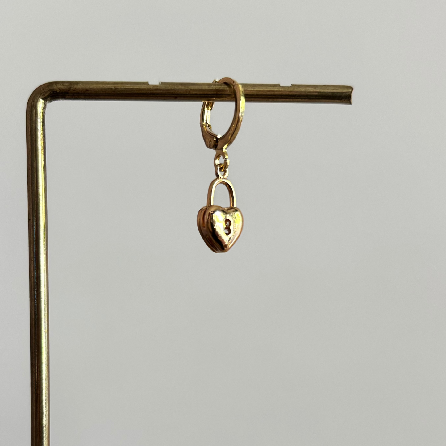 Een goudkleurig klapoorringetje met een hangend hartvormig slotje als bedel, gepresenteerd op een gouden displaystandaard tegen een wit-roze achtergrond.