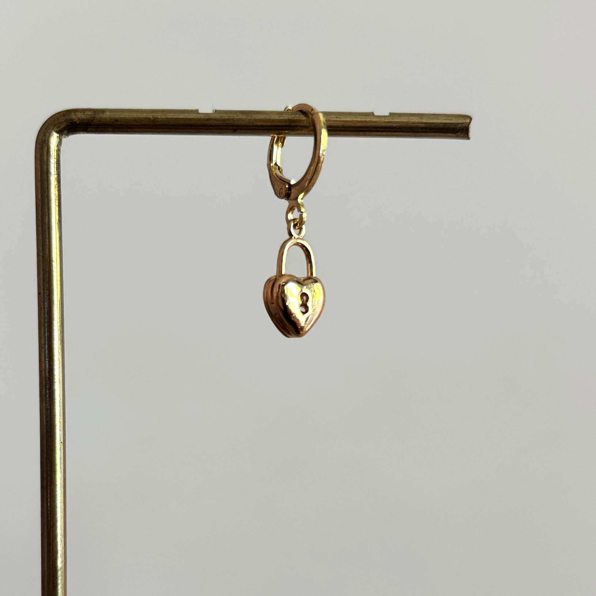 Een goudkleurig klapoorringetje met een hangend hartvormig slotje als bedel, gepresenteerd op een gouden displaystandaard tegen een wit-roze achtergrond.