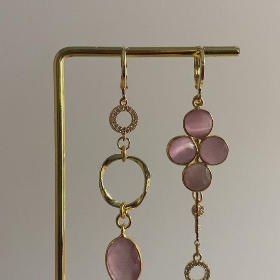 Asymmetrisch oorbellenpaar ‘Lola’: linker oorbel met zirkonia steentje, mini twisted ring en roze glazen bedel; rechter oorbel met taupekleurige steen, kwastje met sleuteltje en zirkonia steentje. Verguld in 14k goud, minimalistisch ontwerp met subtiele details.