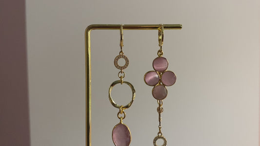 Asymmetrisch oorbellenpaar ‘Lola’: linker oorbel met zirkonia steentje, mini twisted ring en roze glazen bedel; rechter oorbel met taupekleurige steen, kwastje met sleuteltje en zirkonia steentje. Verguld in 14k goud, minimalistisch ontwerp met subtiele details.